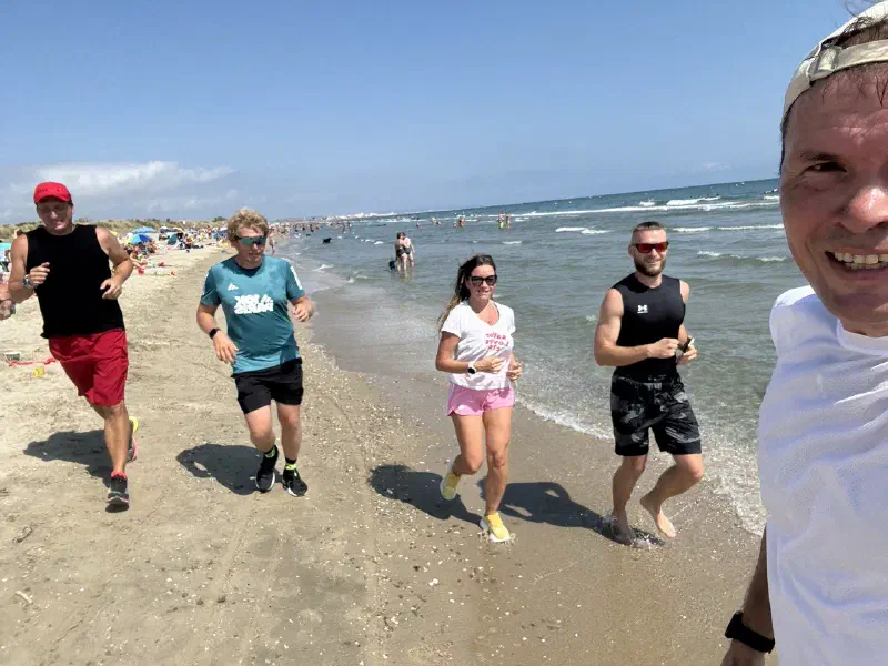 Five people jogging along the water's edge on a sunny day, one of them filming the group as they run