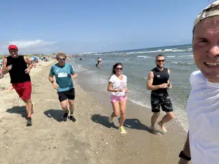 Five people jogging along the water's edge on a sunny day, one of them filming the group as they run