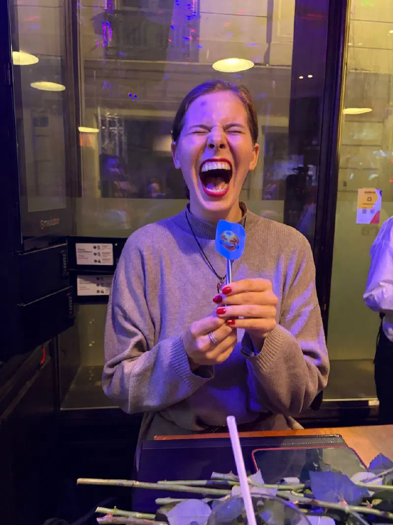 A young woman roaring with laughter, holding a paper emoji prop on a stick; roses lie on the table in front of her