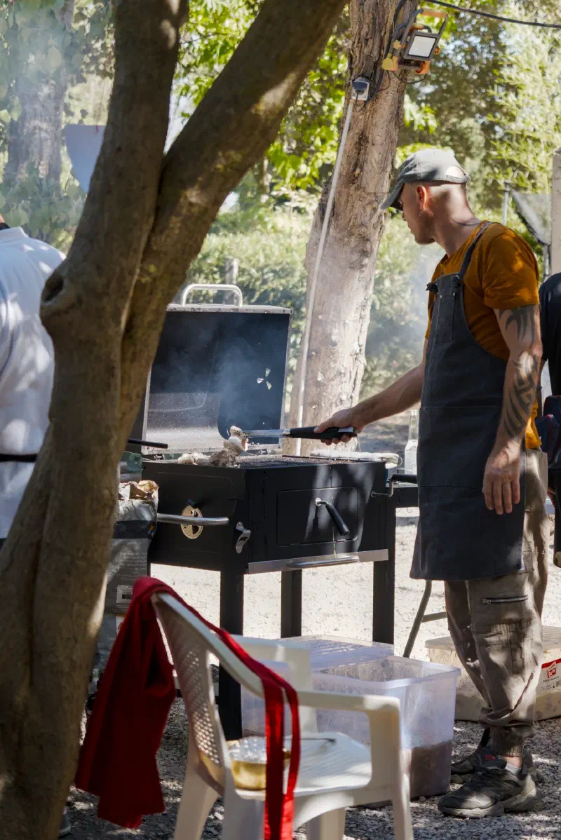 A cook in an apron turns the meat on a black barbecue in the shade of the trees