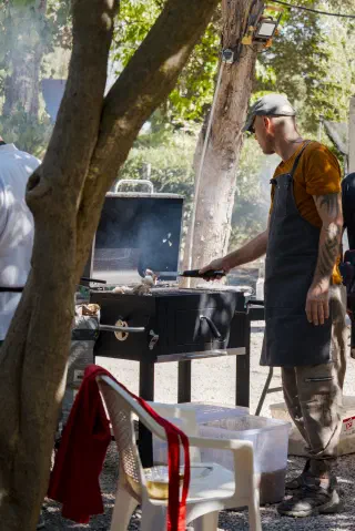A cook in an apron turns the meat on a black barbecue in the shade of the trees