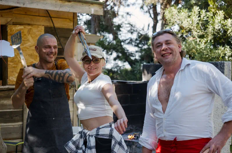 Three people by the summer kitchen with a spatula and tongs, a fire burning in the hearth behind them