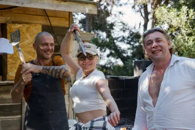 Three people by the summer kitchen with a spatula and tongs, a fire burning in the hearth behind them