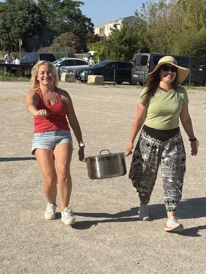 Two smiling women carry a big steel pot across the park grounds