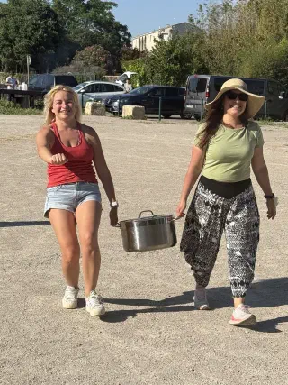 Two smiling women carry a big steel pot across the park grounds