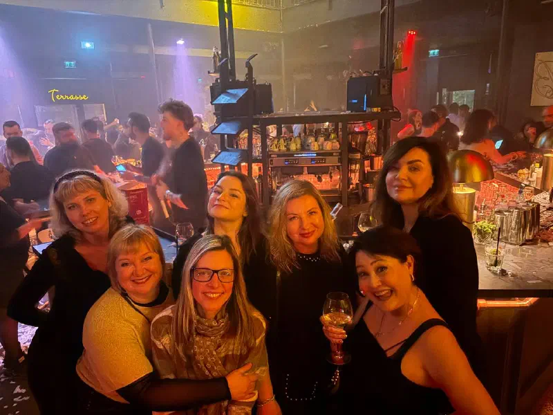 A group of women smiling for the camera in a club, with warm lights, a bar counter and dancers behind.