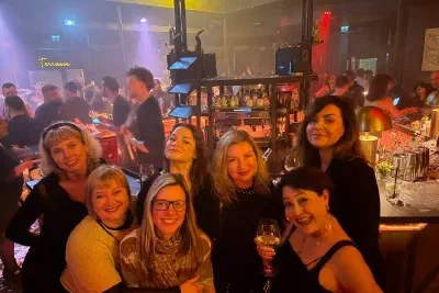 A group of women smiling for the camera in a club, with warm lights, a bar counter and dancers behind.