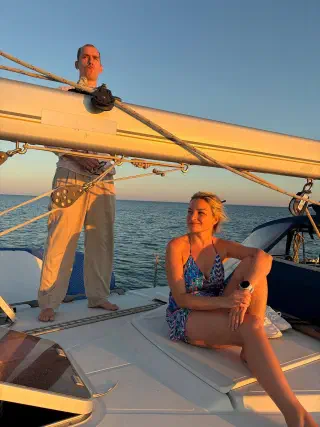 Sunset light on the sailboat: a woman sits on deck under the boom, a participant stands beside her, sea to the horizon
